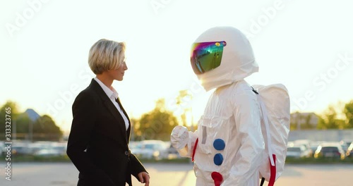 Young Caucasian beautiful woman with short fair hair dancing happily together with an astronaut in the full armor costume at the parking. photo