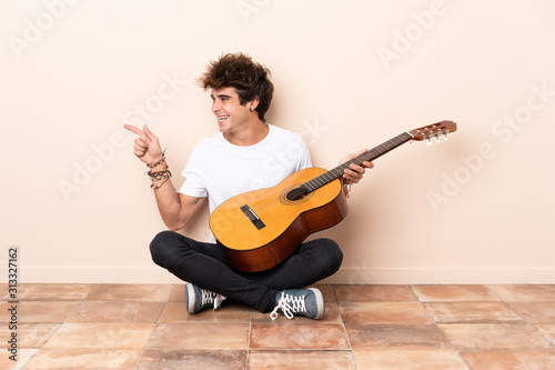 Young caucasian man with a guitar sitting on the floor pointing to the side to present a product