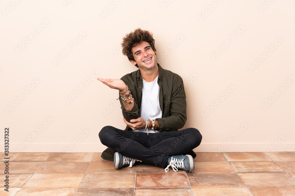Young caucasian man sitting on the floor extending hands to the side for inviting to come Young caucasian man sitting on the floor extending hands to the side for inviting to come