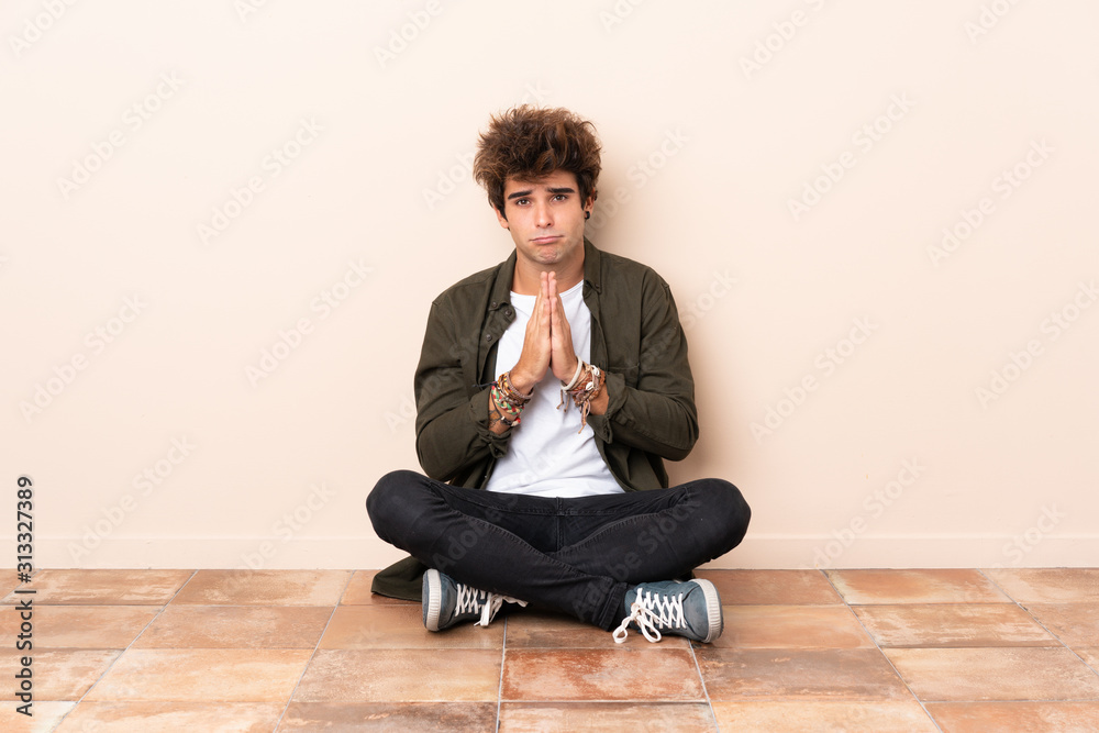 Young caucasian man sitting on the floor keeps palm together. Person asks for something Young caucasian man sitting on the floor keeps palm together. Person asks for something