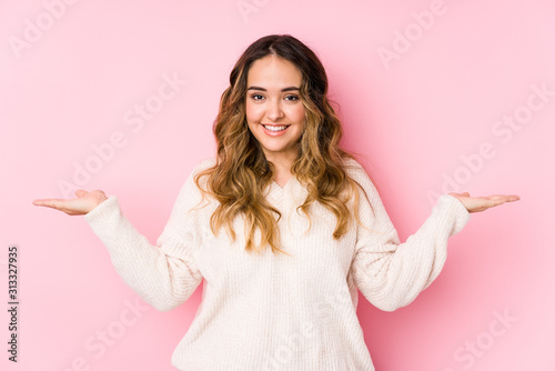 Young curvy woman posing in a pink background isolated makes scale with arms, feels happy and confident.