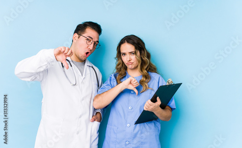 Young doctor couple posing in a blue background isolated showing thumb down and expressing dislike.