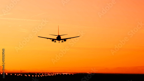 Back view silhouette of airplane landing at the airport at sunny orange and purple sunset. Runway end identification lights and strobe glowing.