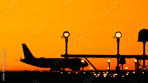 Silhouette of airplanes at the airport at sunny orange and purple sunset. Plane taxiing on the runway preparing for journey. Runway end identification lights and strobe glowing.