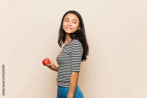 Young asian woman holding an apple looks aside smiling  cheerful and pleasant.