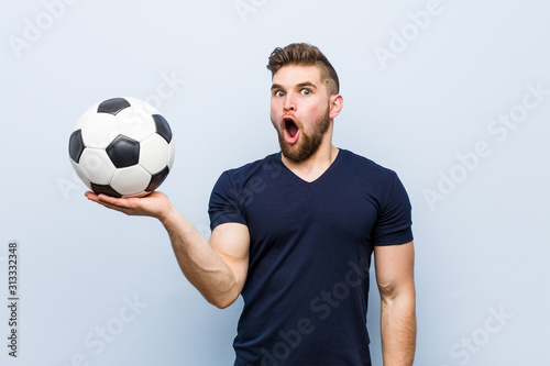 Young caucasian man holding a soccer ball impressed holding copy space on palm.
