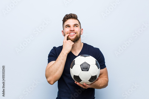 Young caucasian man holding a soccer ball relaxed thinking about something looking at a copy space.