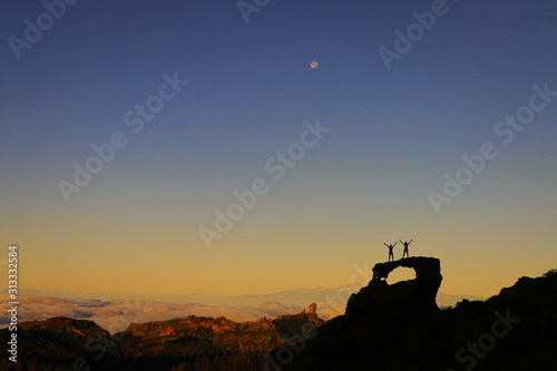 Two silhouettes victoriously standing on a rock arch on a mountain top with clouds below during yellow sunrise. Roque Nublo and Teide mountain in the background. Moon on the blue sky. Canary Islands