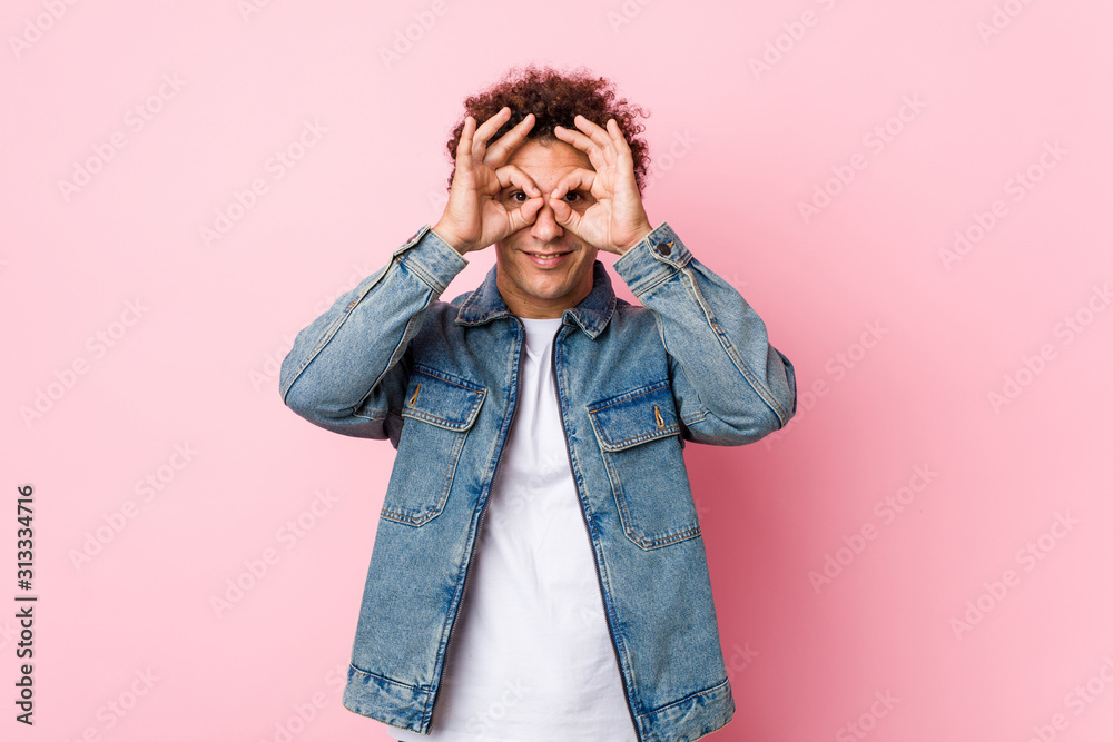 Curly mature man wearing a denim jacket against pink background showing okay sign over eyes Curly mature man wearing a denim jacket against pink background showing okay sign over eyes