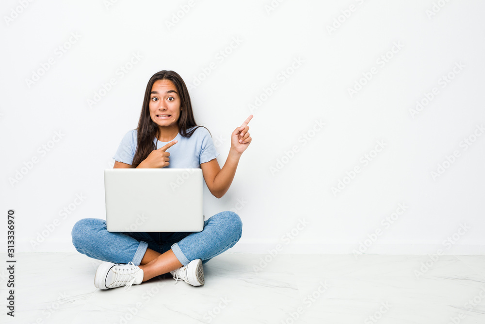 Young mixed race indian woman sitting working on laptop shocked pointing with index fingers to a copy space. Young mixed race indian woman sitting working on laptop shocked pointing with index fingers to a copy space.