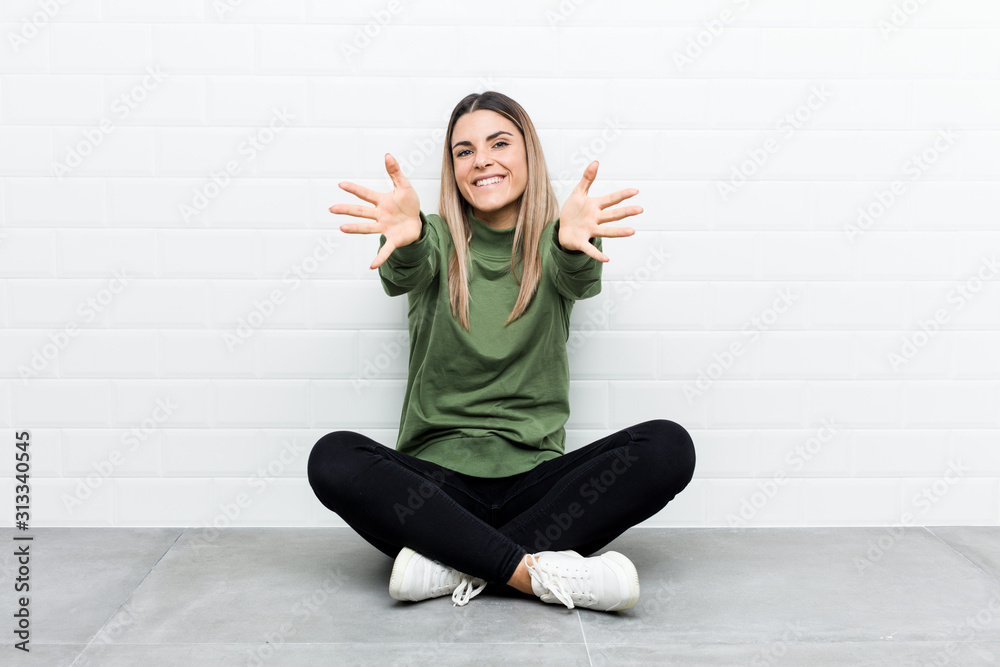 Young caucasian woman sitting on the floor feels confident giving a hug to the camera.