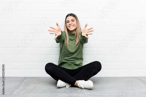 Young caucasian woman sitting on the floor feels confident giving a hug to the camera.