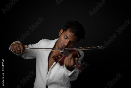 Portrait of young and attractive Indian Bengali brunette man with white western suit and hat is playing violin in front of black studio copy space background. Indian fashion portrait and lifestyle.