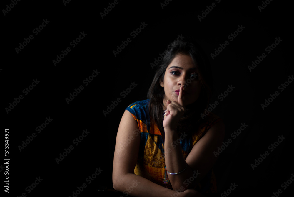 Fashion portrait of young and attractive Indian Bengali brunette girl with colorful western dress with fingers on her lips in front of a black studio background. Indian fashion portrait and lifestyle.