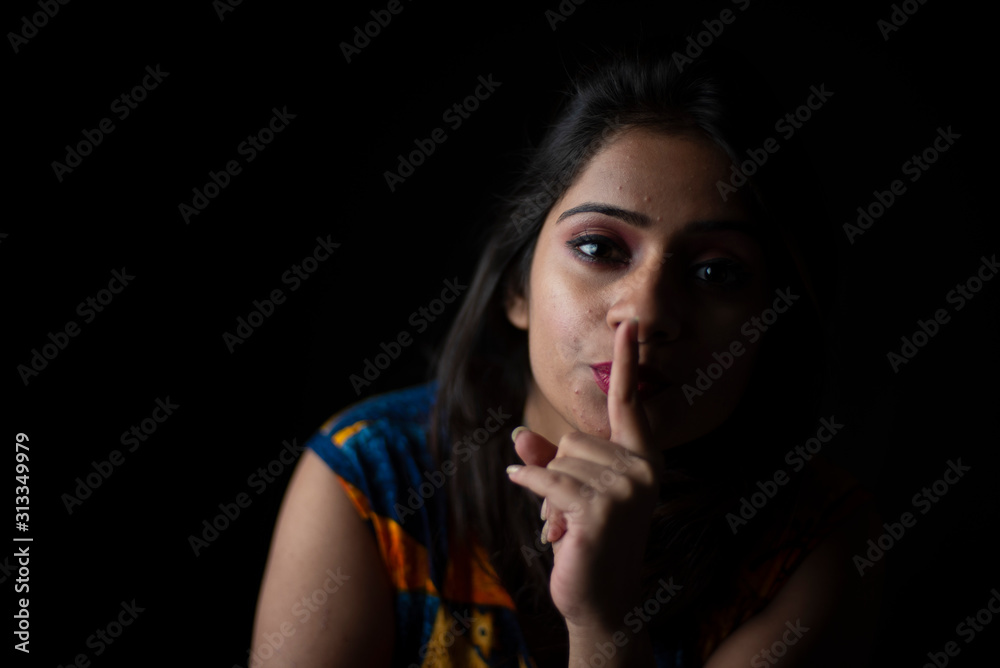 Fashion portrait of young and attractive Indian Bengali brunette girl with colorful western dress with fingers on her lips in front of a black studio background. Indian fashion portrait and lifestyle.