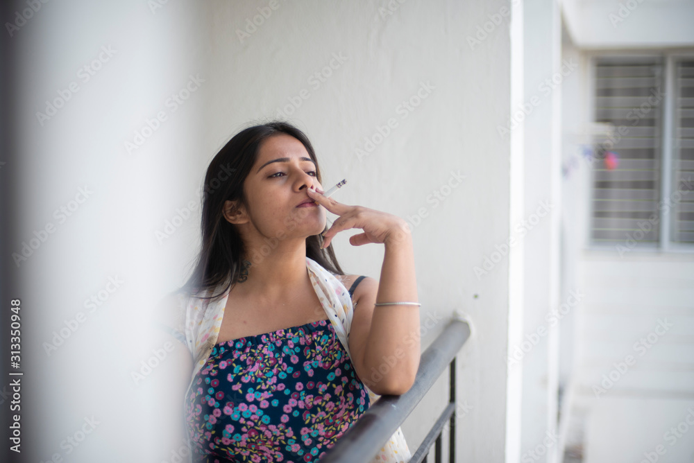 An young and attractive Indian Bengali brunette woman in white sleeping wear is smoking cigarette on the balcony in the morning in white background. Indian lifestyle. An young and attractive Indian Bengali brunette woman in white sleeping wear is smoking cigarette on the balcony in the morning in white background. Indian lifestyle.