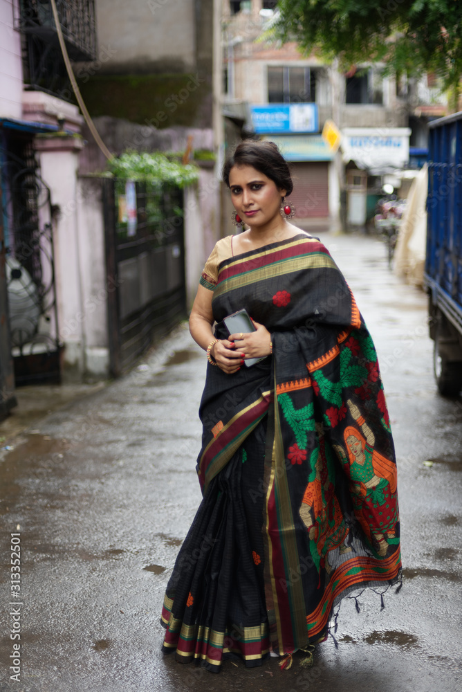 Portrait of a beautiful brunette Indian bengali thoughtful woman in traditional wear sari standing in a lane on a drenched morning of Durga Puja festival in urban  background. Indian lifestyle.