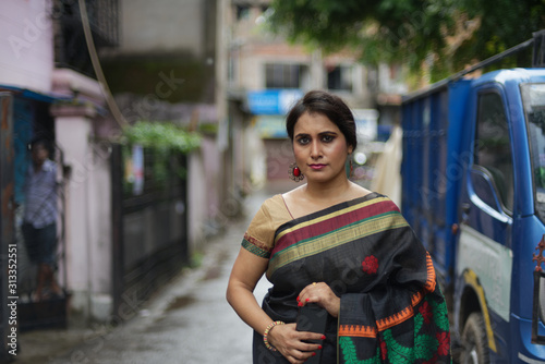 Portrait of a beautiful brunette Indian bengali thoughtful woman in traditional wear sari standing in a lane on a drenched morning of Durga Puja festival in urban background. Indian lifestyle.