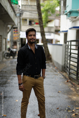 Portrait of a handsome brunette Indian bengali smart man in shirt and trousers standing in a drenched street in the morning of Durga Puja festival in urban background. Indian lifestyle.