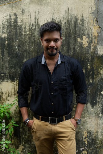 Portrait of a handsome brunette Indian bengali smart man in black shirt and trousers standing beside a tree in the morning of Durga Puja festival in textured background. Indian lifestyle. © abir