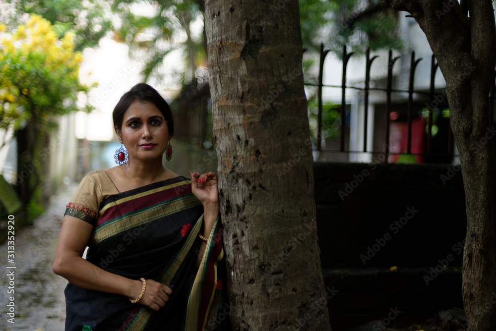 Naklejka premium Portrait of a beautiful brunette Indian bengali thoughtful woman in traditional wear sari standing in a lane on a drenched morning of Durga Puja festival in urban background. Indian lifestyle.