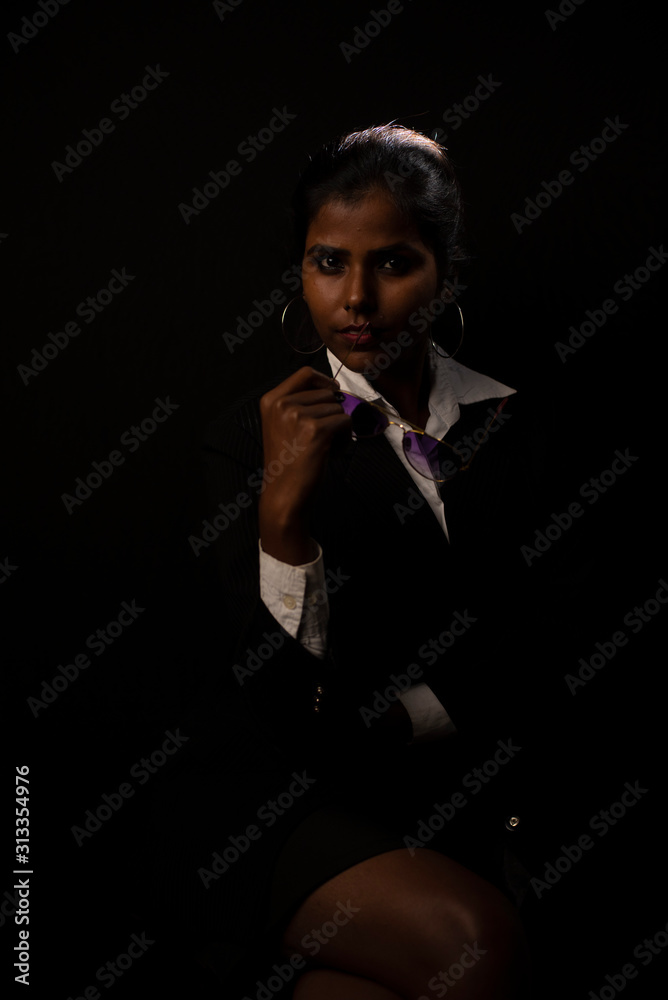 Fototapeta premium An young and attractive dark skinned Indian Bengali brunette woman in western jacket, skirt and goggles sitting under spotlight in a black copy space background. Indian fashion photography.