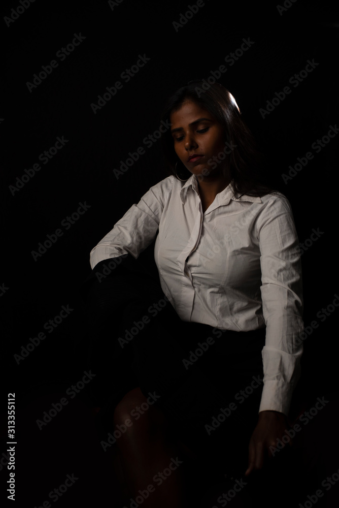 Young and attractive dark skinned Indian Bengali brunette woman in western shirt and skirt with jacket on shoulder under spotlight in black studio copy space background. Indian fashion photography.