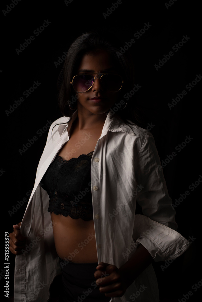 Fashion portrait of young and attractive dark skinned Indian Bengali brunette woman in western lingerie and glasses removing white shirt  in black copy space background. Indian fashion photography.