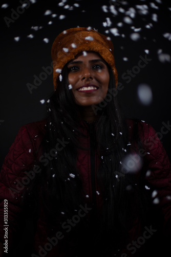 Portrait of an young and attractive dark skinned Indian woman wearing woolen cap and jacket playing with snowflakes in a snowy evening in dark background. Winter and Christmas