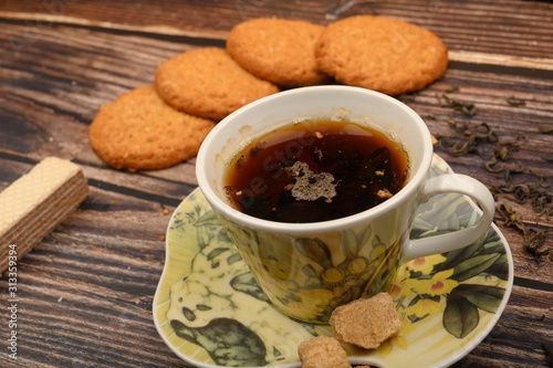 A Cup of black tea, tea leaves, pieces of brown sugar, oatmeal cookies, waffles on a wooden background. Close up.