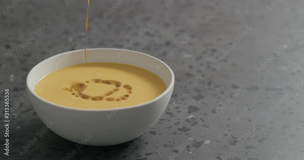 Slow motion pour olive oil with spices to pumpkin cream soup in white bowl on terrazzo countertop Slow motion pour olive oil with spices to pumpkin cream soup in white bowl on terrazzo countertop