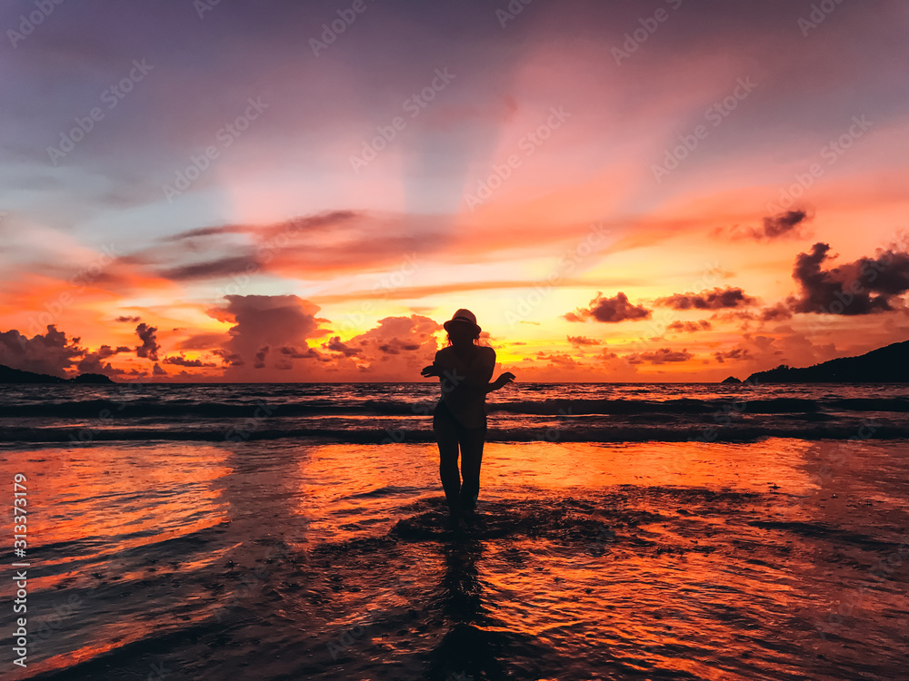 A beautiful young woman is splashing water in a hat is dancing on the seacoast on the background of sunset. Sense of freedom. Amazing mesmerizing landscape