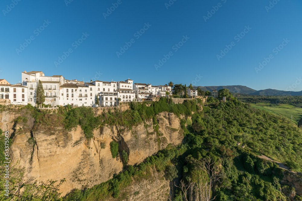 ronda village at the edge of cliffside with trees and white houses against sky, Andalusia, Spain