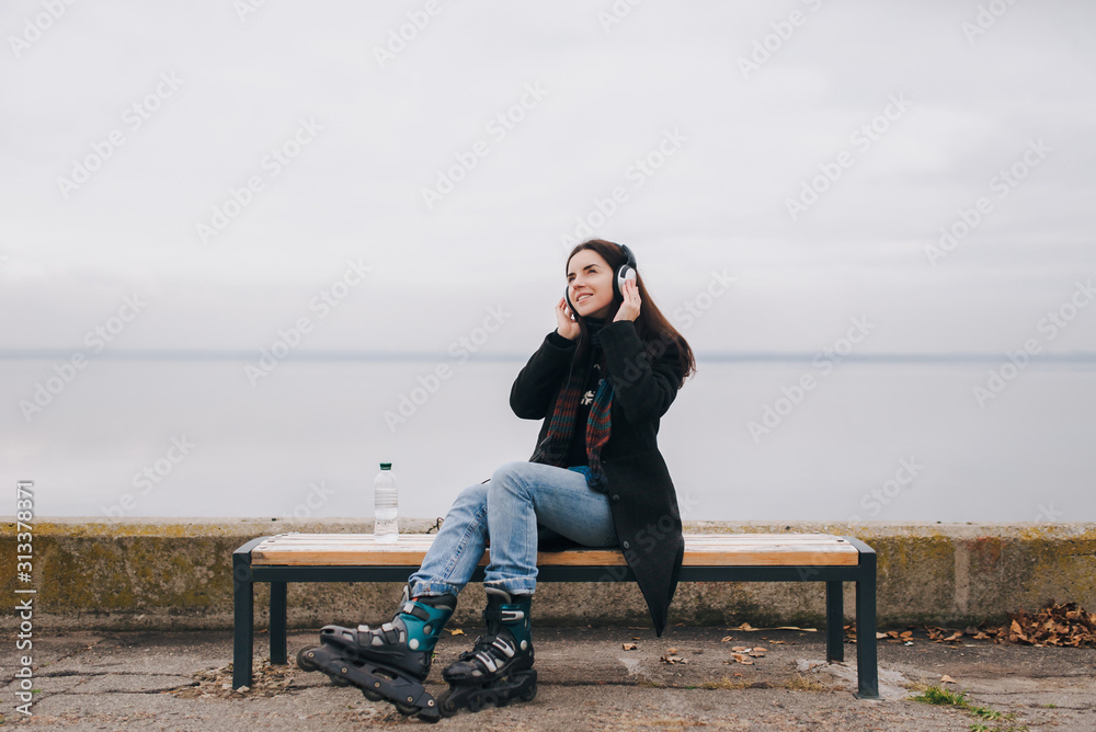 A beautiful caucasian girl in roller skates is listening to music with headphones. The girl enjoys the music and smiles.