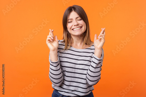 Portrait of hopeful joyous young woman with brown hair in long sleeve shirt standing  raising finger crossed while making wish  confident to win. indoor studio shot isolated on orange background