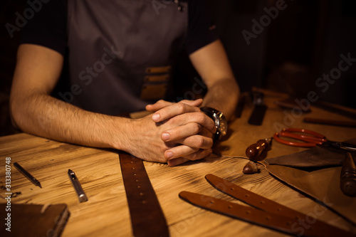 Working process of the leather belt in the leather workshop. Man holding hands on wooden table. Crafting tools on background. Tanner in old tannery. Close up men arm. Interlock fingers.