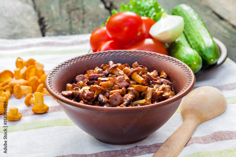 Fototapeta premium Fried chanterelles with onion in rustic bowl and plate with fresh vegetables for salad on background