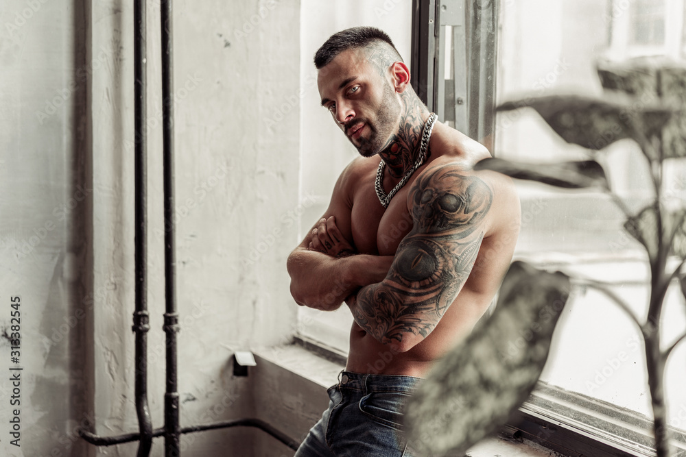 Close up portrait of Sexy naked male model with tattoo and magic eyes  standing in hot pose on near the window. Loft room interior with grey  concrete wall. Professional Studio image. Stock