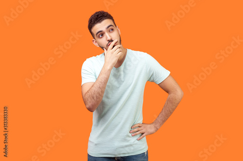 Need to think. Portrait of thoughtful brunette man with beard in white t-shirt holding his chin and pondering idea, confused not sure about solution. indoor studio shot isolated on orange background