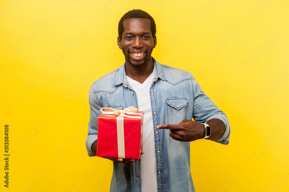 Portrait of joyous handsome man in denim casual shirt smiling at camera and pointing at red gift box, satisfied with present, enjoying holidays. indoor studio shot isolated on yellow background Portrait of joyous handsome man in denim casual shirt smiling at camera and pointing at red gift box, satisfied with present, enjoying holidays. indoor studio shot isolated on yellow background