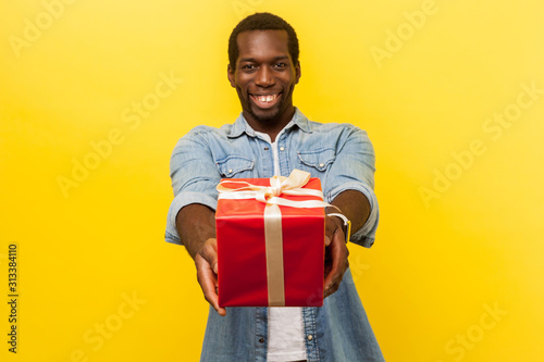 Present for you! ! Portrait of positive generous man in denim casual shirt giving red gift box and smiling at camera, congratulating on holiday. indoor studio shot isolated on yellow background © khosrork