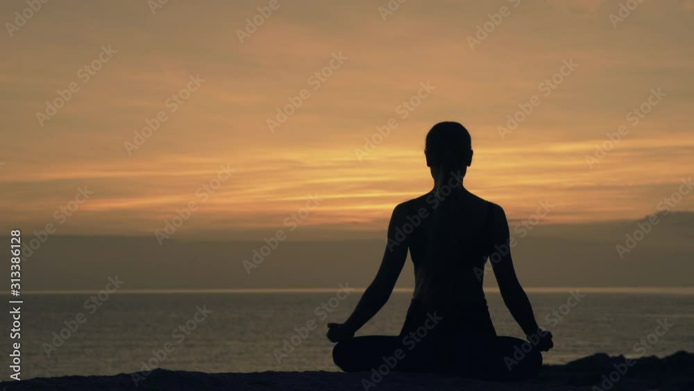 Silhouette of a meditating young woman sitting in lotus position on a rock by the sea against the background of dawn Silhouette of a meditating young woman sitting in lotus position on a rock by the sea against the background of dawn