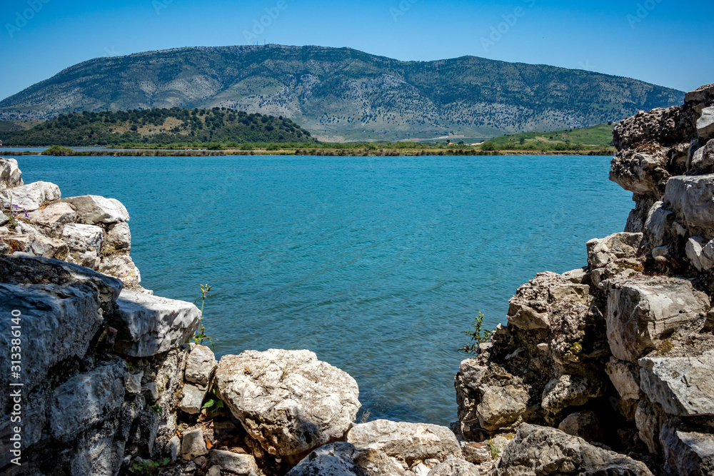 Fototapeta premium Sunny spring day view of salt lagoon lake, valley and mountain in the national park of Butrint in Albania, Europe. Fortress stone ruined wall in the foreground.