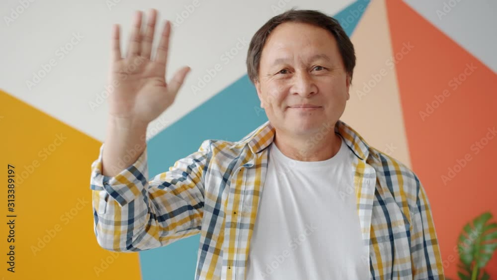 Cheerful Asian man is waving hand smiling standing indoors at home alone being friendly. Interiors, joyful mature people and human emotions concept. Cheerful Asian man is waving hand smiling standing indoors at home alone being friendly. Interiors, joyful mature people and human emotions concept.
