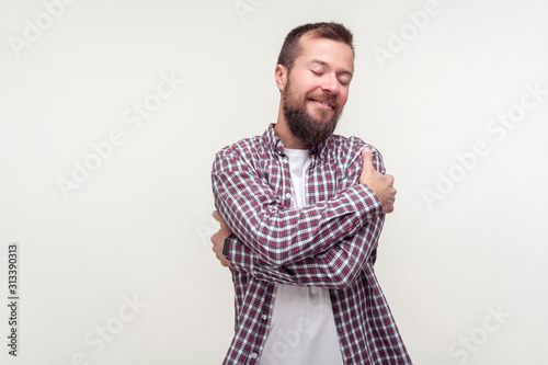 Love yourself. Portrait of smiling confident bearded man in plaid shirt embracing himself with satisfied pleased expression, positive self-esteem. indoor studio shot isolated on white background