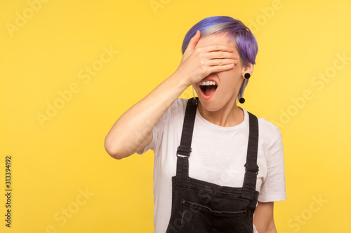 Don't want to look. Portrait of scared shocked girl with violet hair in denim overalls covering eyes with hand, expressing dissatisfaction with what she sees, ignoring shameful content. studio shot