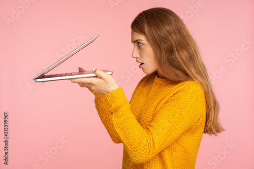 Obraz na plátně Portrait of curious ginger girl in sweater carefully peeking into half-opened laptop and expressing surprise amazement, spying on forbidden content