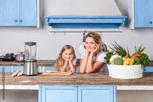 Young cheerful mother and little daughter leaning on table, standing in kitchen with modern furniture, basket of fresh vegetables on table, eco-friendly vegetarian food, vegan nutrition, healthcare