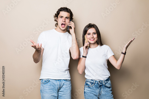 Portrait of a happy young couple talking on mobile phones sitting together over beige background,
