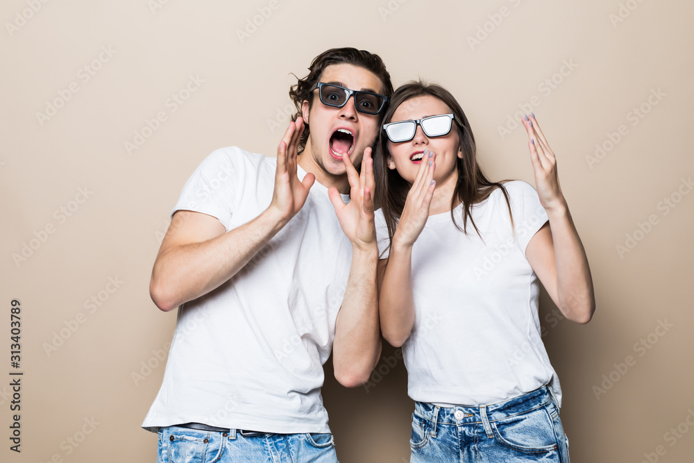Two young shocked lovers are amazed with wide open eyes, mouthes, fixing sunglasses, in a casual wear on beige background Two young shocked lovers are amazed with wide open eyes, mouthes, fixing sunglasses, in a casual wear on beige background